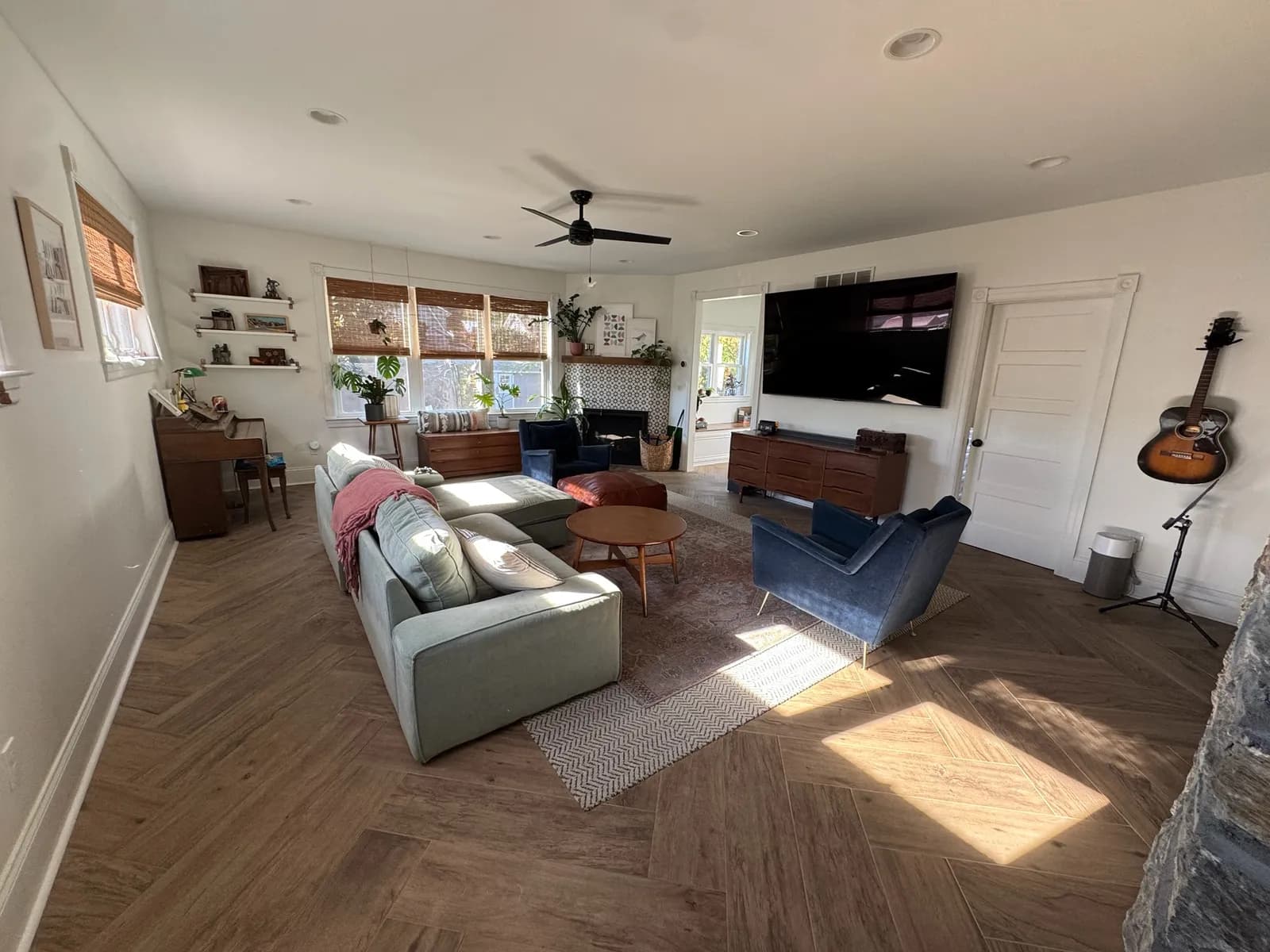Family room in the Glenside addition: herringbone plank floor, a sectional and blue armchairs, a wall-mounted TV, a ceiling fan, and windows along the far wall