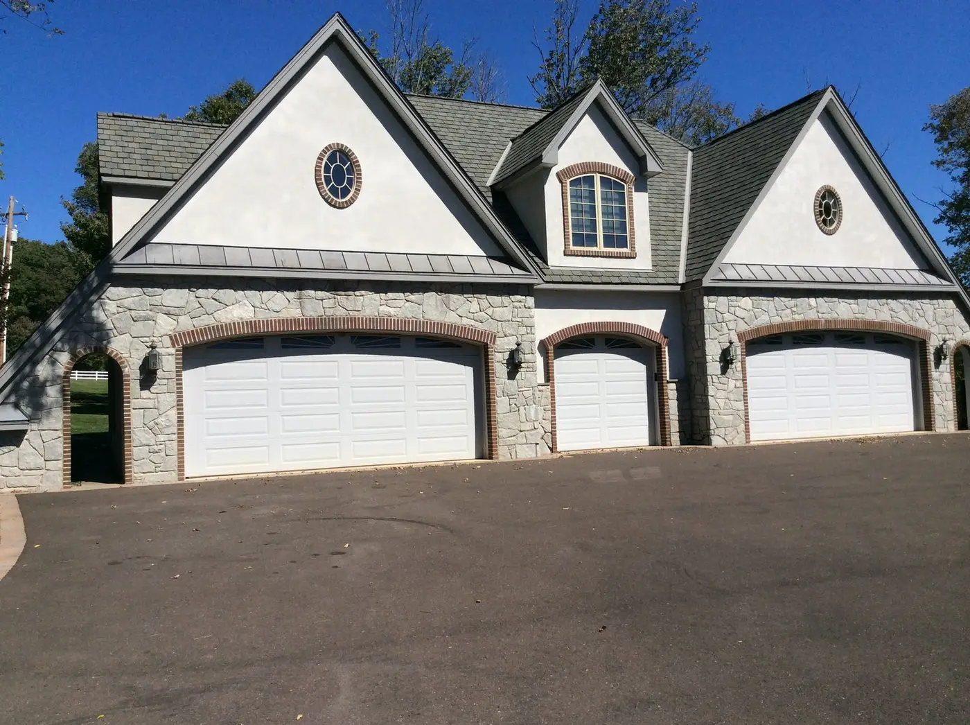 Three-bay garage with white doors, stone and stucco walls, and dormers above, at the Hilltown, PA custom home