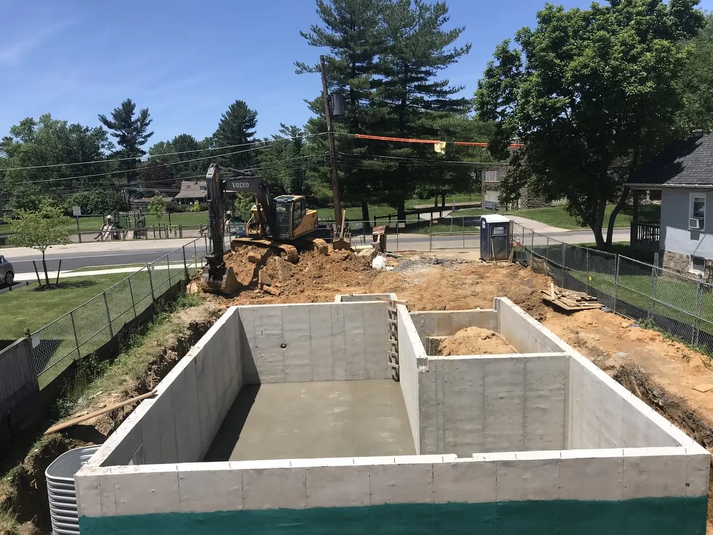 Poured concrete foundation walls in the open excavation, with an excavator and the street behind