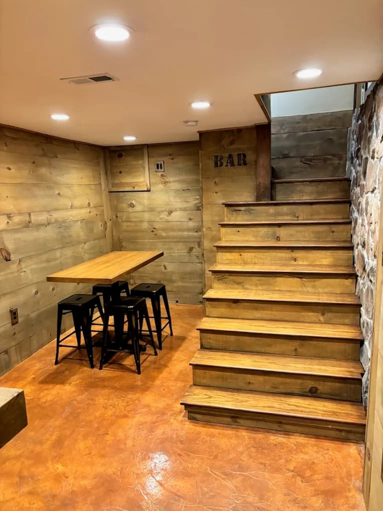 Finished basement with wood-plank walls, a stained concrete floor, a bar-height table with black stools, and wood stairs beside a stone wall