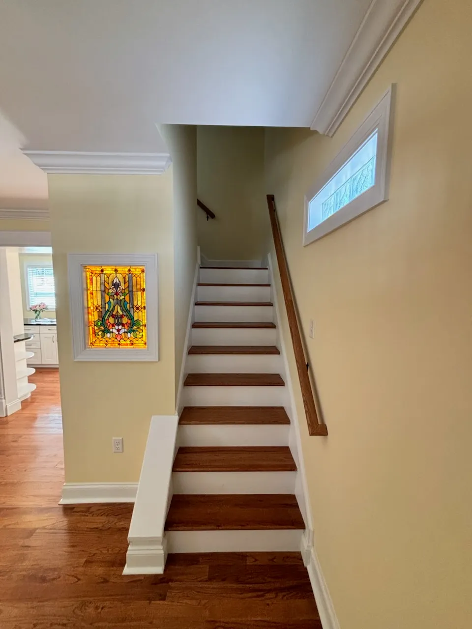Stair with wood treads and white risers rising from a hardwood-floored hall, with a stained glass panel on the wall beside it
